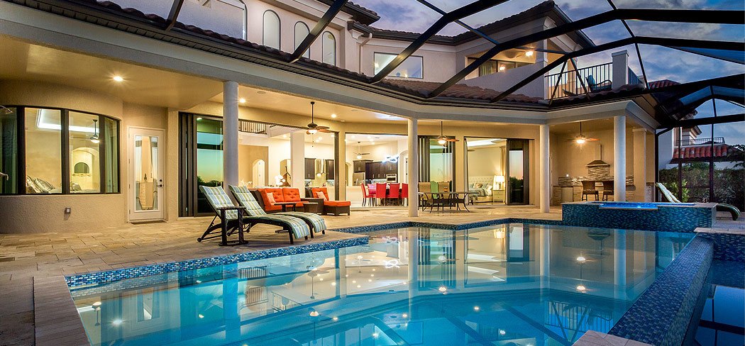 Screened pool and spa behind a two-story home at dusk, with an outdoor kitchen under the lanai
